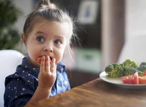 A young girl eating fresh tomatoes and brocoli while sitting at a table.